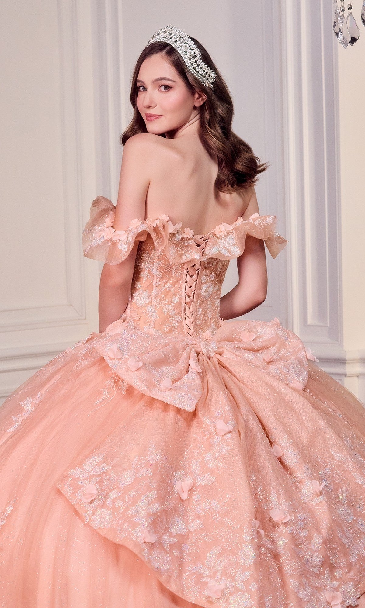 A young woman poses with her back to the camera in an elegant room, wearing the Quinceanera Dress 15731 By Ladivine—an off-the-shoulder peach ball gown with lace, 3D flowers, and a tiara.